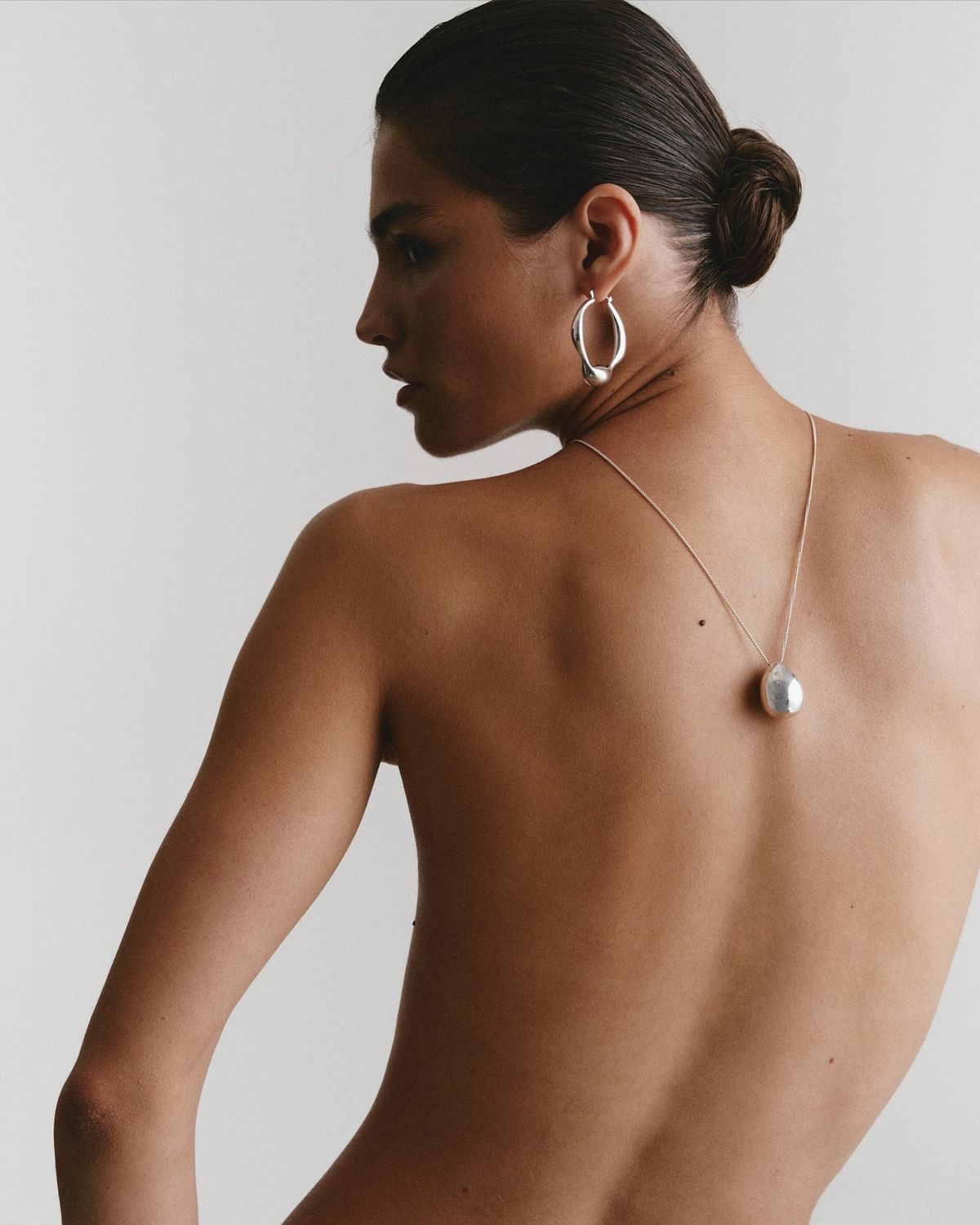Woman with her hair in a bun, showing her bare back, wearing a silver necklace and an abstract silver earring posing for TANNED TO - an automated spray tan studio in Toronto.