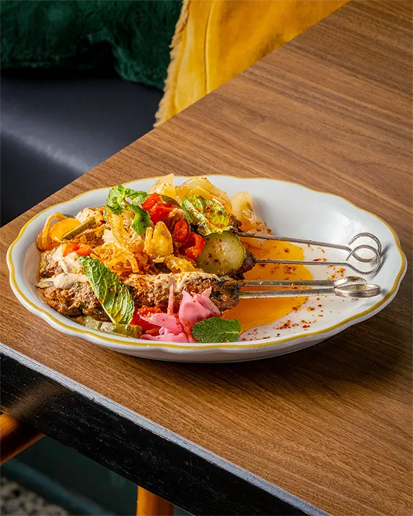 Brochettes de kafta grillées garnies de légumes frais, oignons frits, et herbes, servies sur une assiette blanche sur une table en bois.