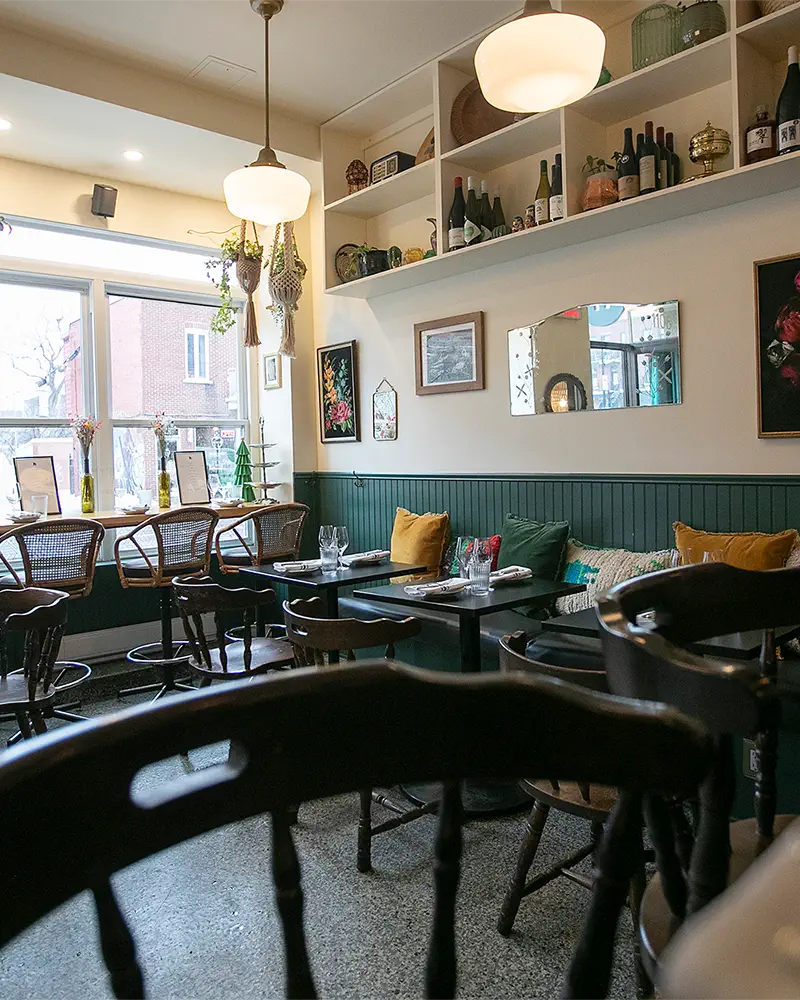 Intérieur lumineux d'un bistro avec des tables dressées, chaises en bois, étagères murales avec bouteilles et décorations, et grandes fenêtres laissant entrer la lumière naturelle.