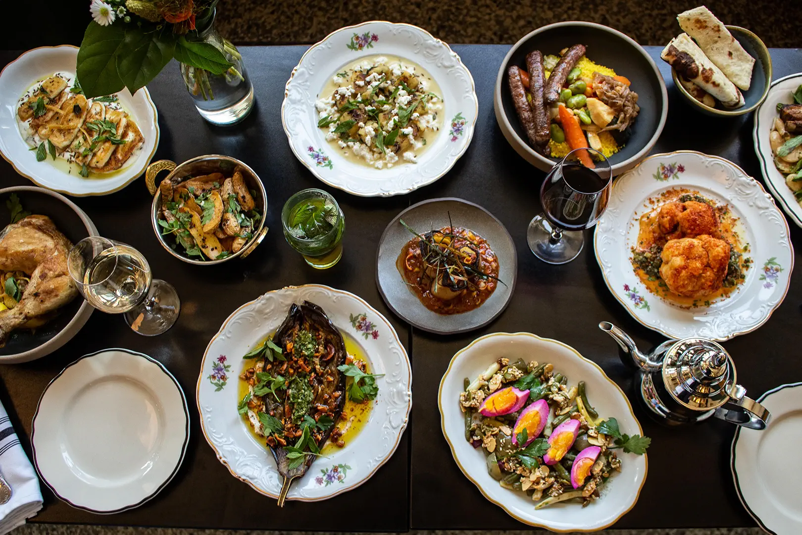 Table avec divers plats marocains, y compris légumes grillés, viande, salades, et boissons comme thé et vin rouge.