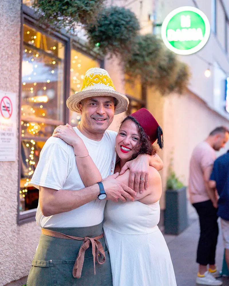Un homme avec un chapeau tissé et un tablier vert serre dans ses bras une femme souriante portant un fez rouge, devant un restaurant avec des lumières décoratives.