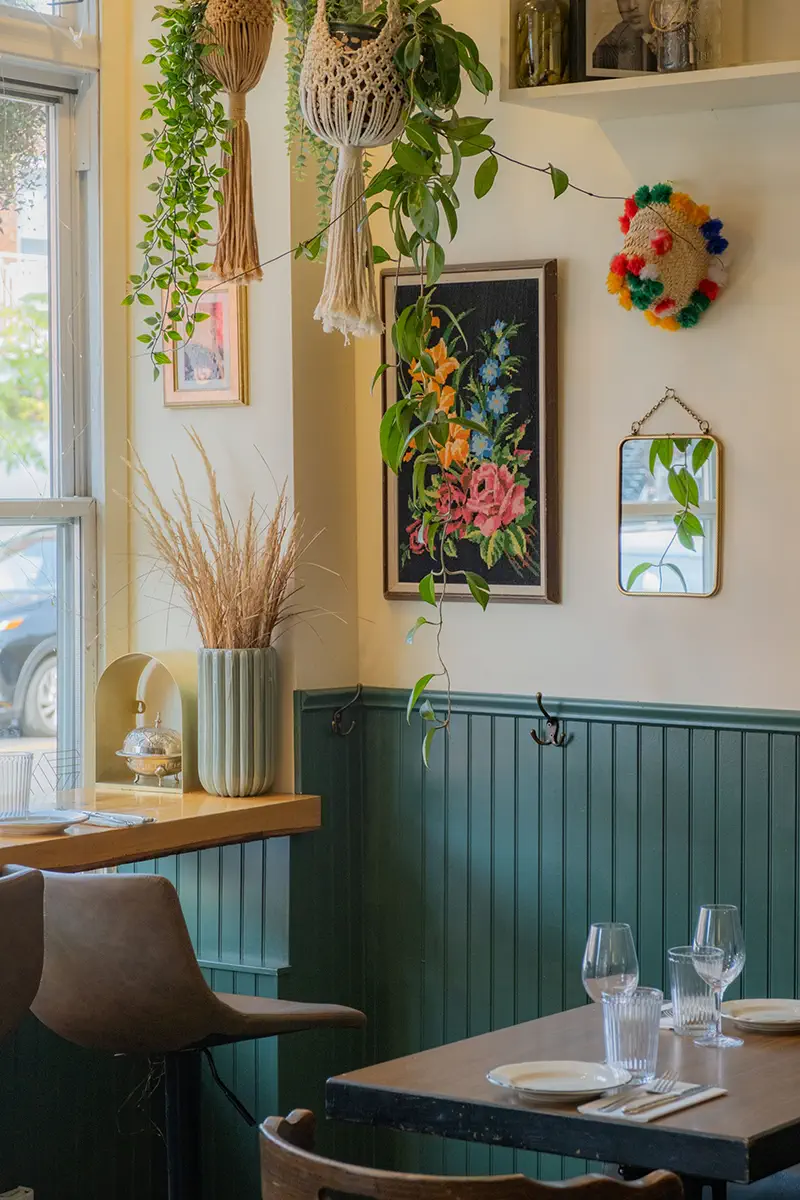 Coin d'un bistro avec chaise en cuir, table dressée, plantes suspendues, vase avec herbes séchées et décorations murales colorées.