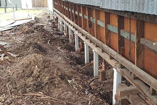 Construction site showing the side of a building lifted on concrete piers with exposed foundation and soil dug out underneath.