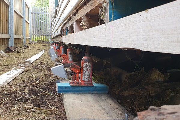 Foundation of a wooden building elevated on red jack stands placed on wooden planks over soil.
