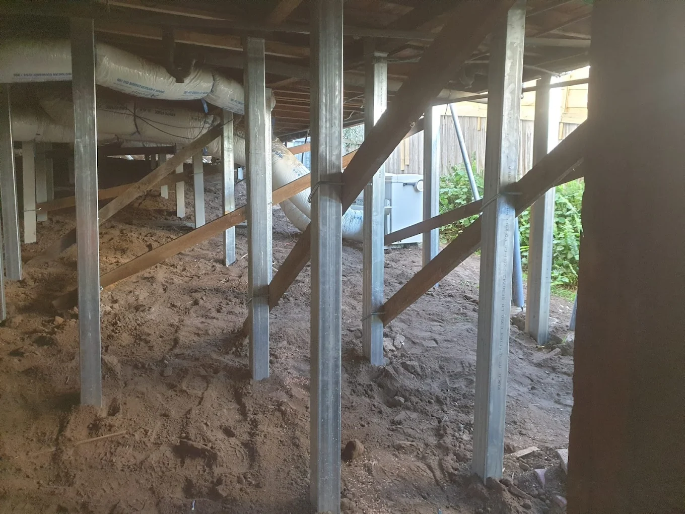 Underneath a raised building showing metal support columns with diagonal wooden braces and exposed ductwork wrapped in insulation.