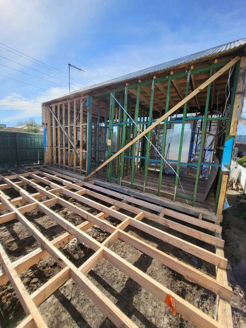 Partially constructed wooden structure with exposed framing and floor joists on bare ground under a blue sky.