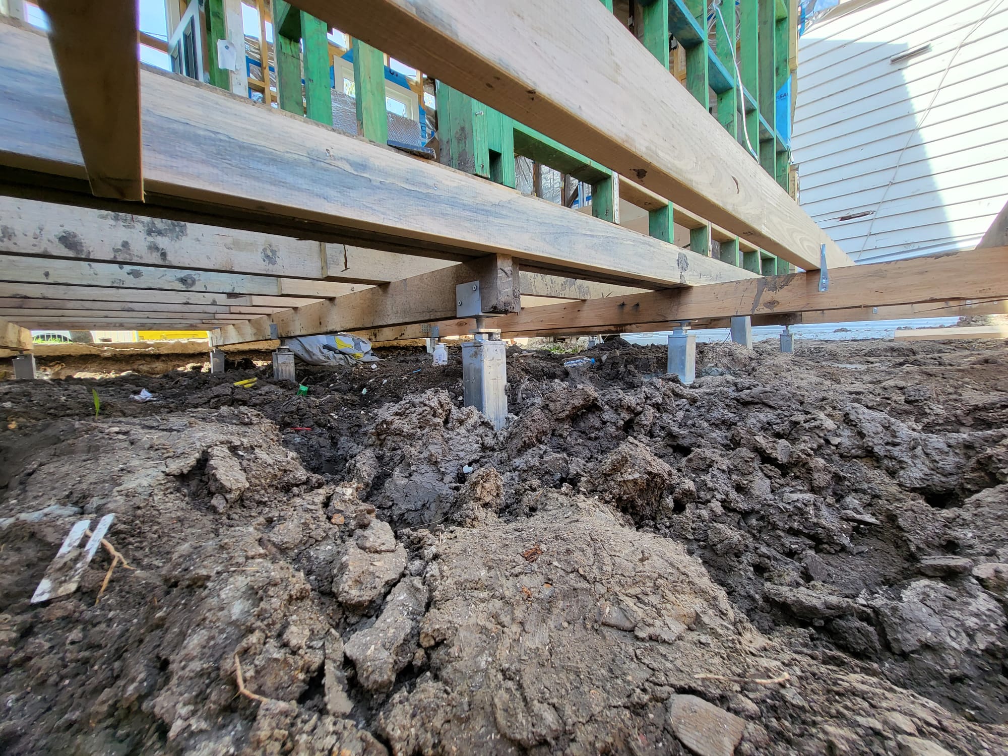 View underneath a wooden house framework showing metal pier foundations embedded in the soil.