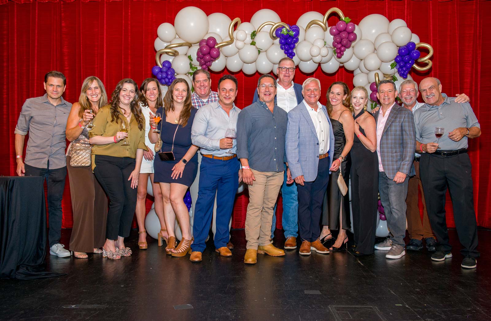 Group of fifteen adults posing happily in front of a red curtain and a white balloon arch decorated with purple and pink grapes, many holding drinks.