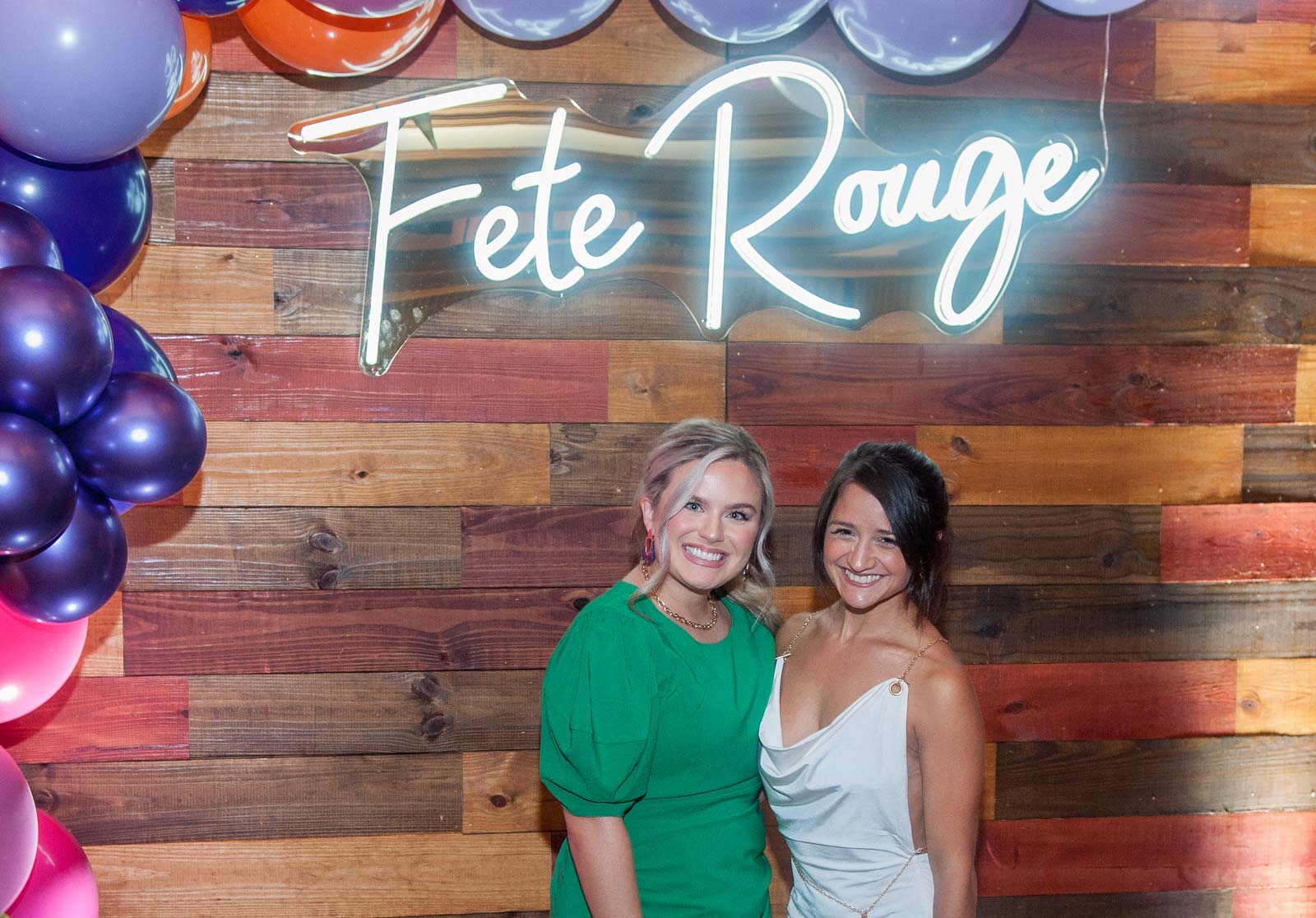 Two smiling women posing in front of a wooden wall decorated with balloons and a glowing neon sign reading 'Fete Rouge'.