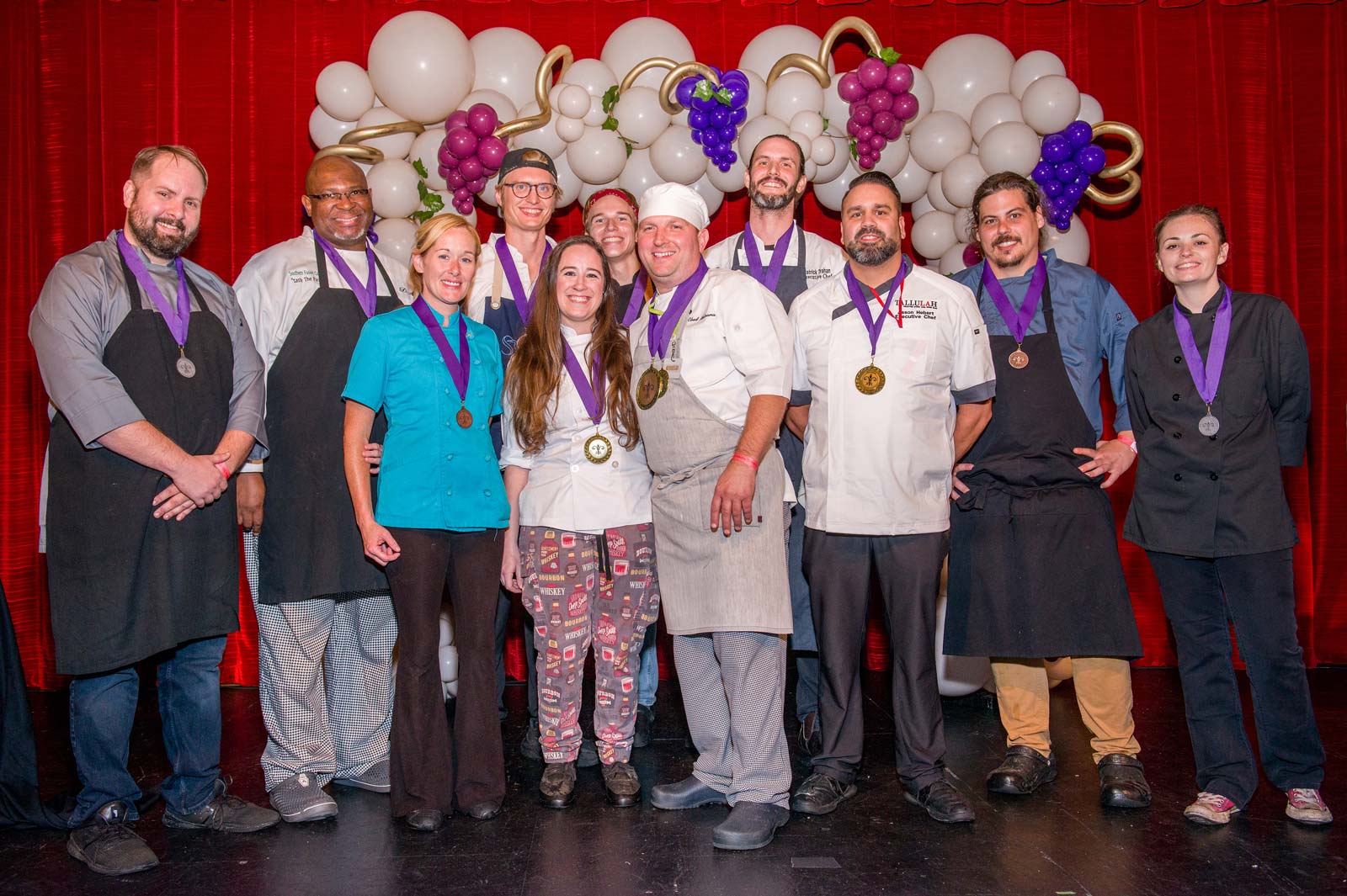 Group of twelve chefs and culinary professionals wearing medals, standing in front of a white balloon arch with grape clusters and a red curtain backdrop.