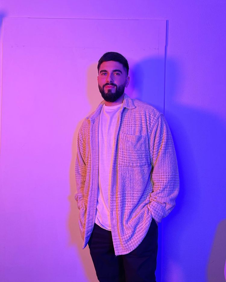 Bearded man wearing a white t-shirt and textured jacket standing against a plain wall lit with purple and blue lighting.