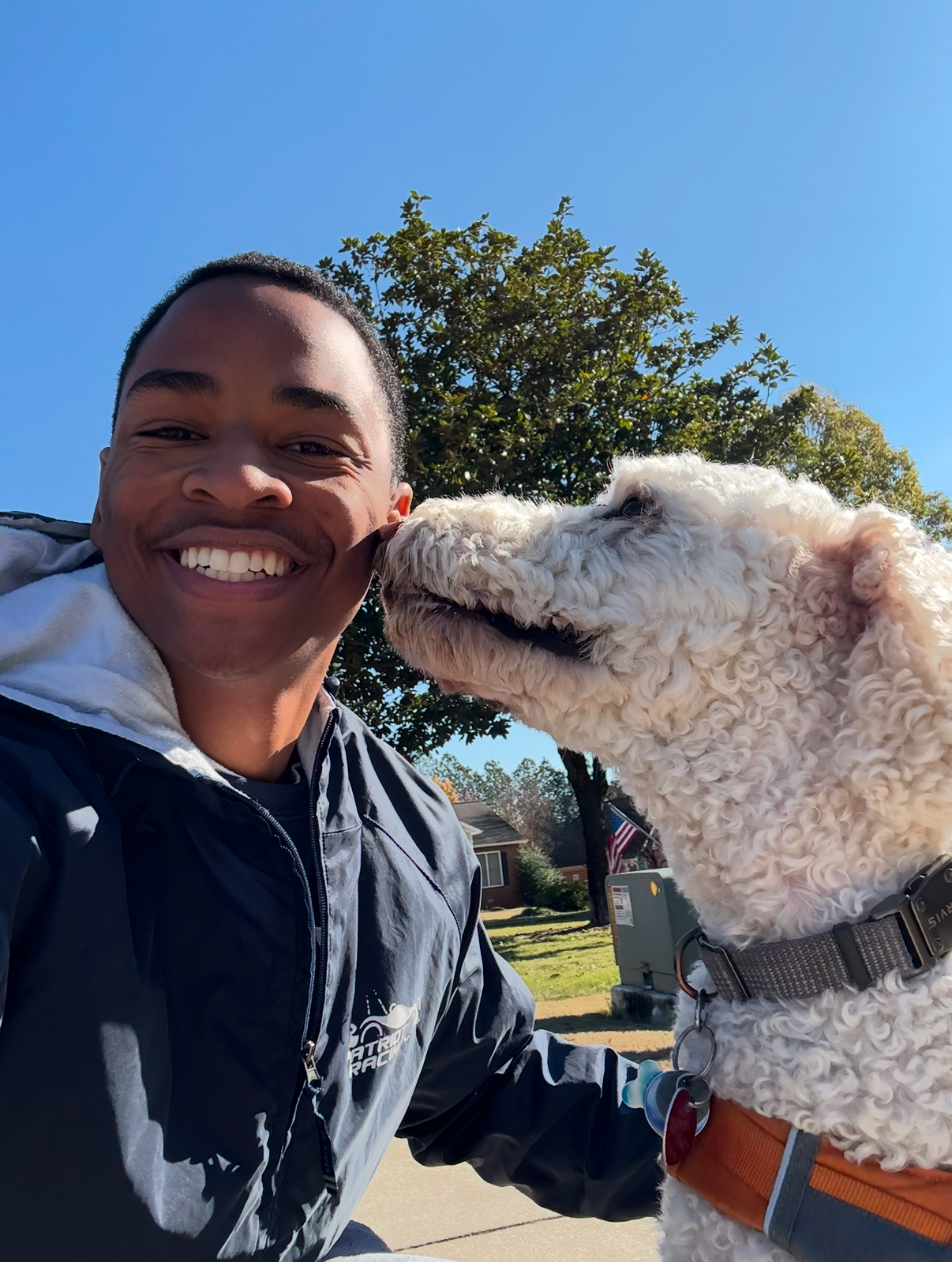 Smiling man outdoors with a white curly dog licking his cheek under clear blue sky.