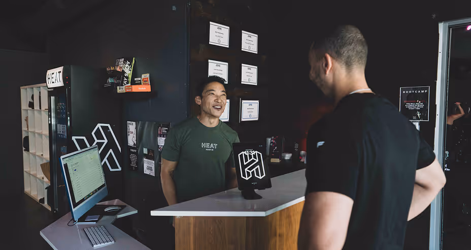 Two men talking at a gym reception desk with certificates on the wall and a computer on a side table.