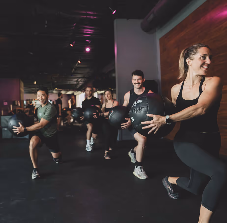 Group of five people in athletic wear performing lunges while holding medicine balls in a gym with dim lighting.