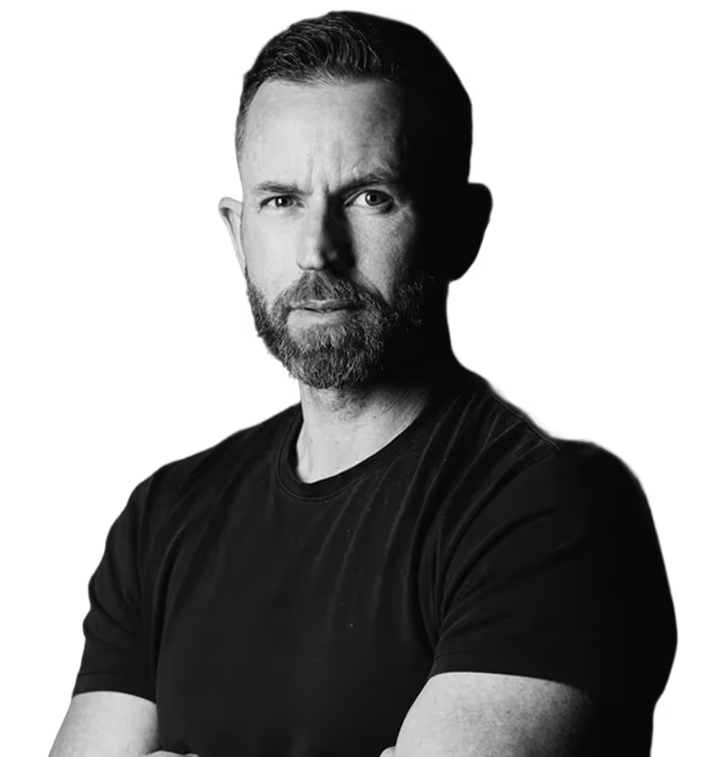 Black and white portrait of a serious bearded man in a black t-shirt with arms crossed against a dark background.