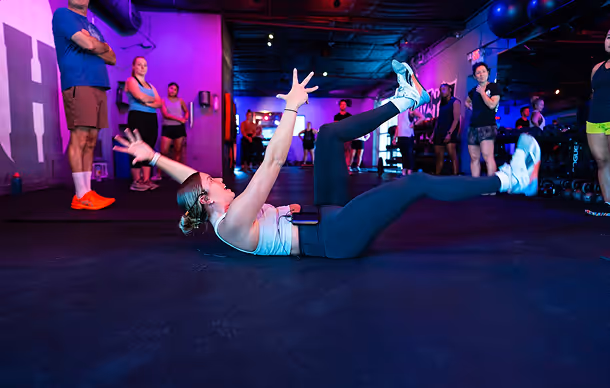 Woman exercising on gym floor with legs raised while others watch in a dimly lit workout space with purple and blue lighting.