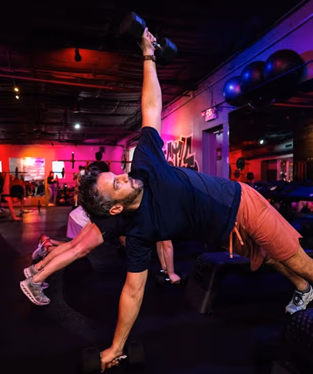 Man balancing in a side plank position while holding dumbbells in a dimly lit gym with a red and purple neon ambiance.