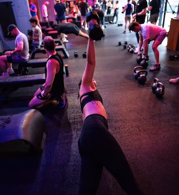 People working out with dumbbells and kettlebells in a busy gym with dim, colorful lighting.
