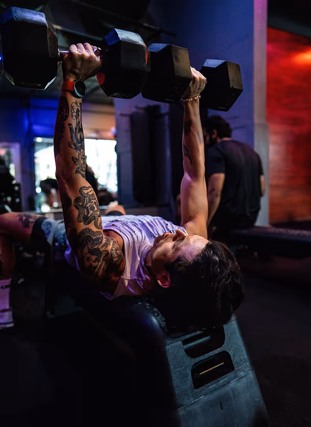 Tattooed man lifting dumbbells while lying on a workout bench in a dimly lit gym.