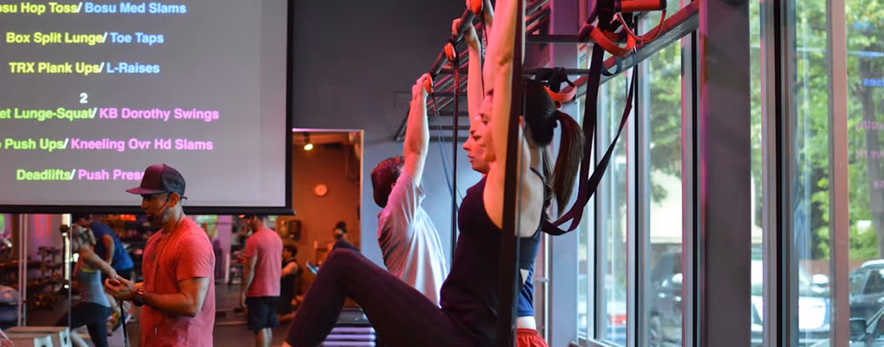 People exercising in a gym using suspension straps attached to a rig near large windows with workout routine projected on a screen.