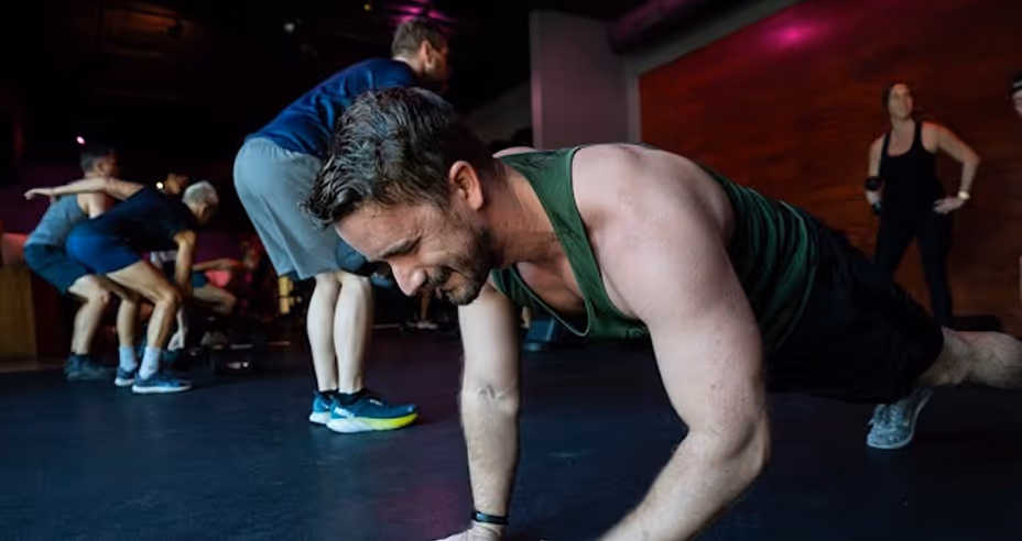 A group fitness class in a gym with several people exercising, focusing on a man in a green tank top doing a push-up.