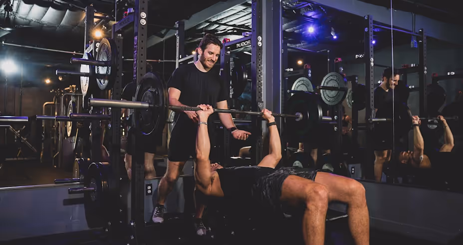 Man lifting a barbell on a bench press with a trainer spotting him in a gym with mirrored walls.