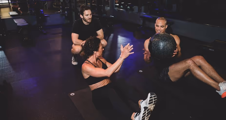 Two people sitting on the floor in a gym passing a medicine ball, with a trainer watching them.