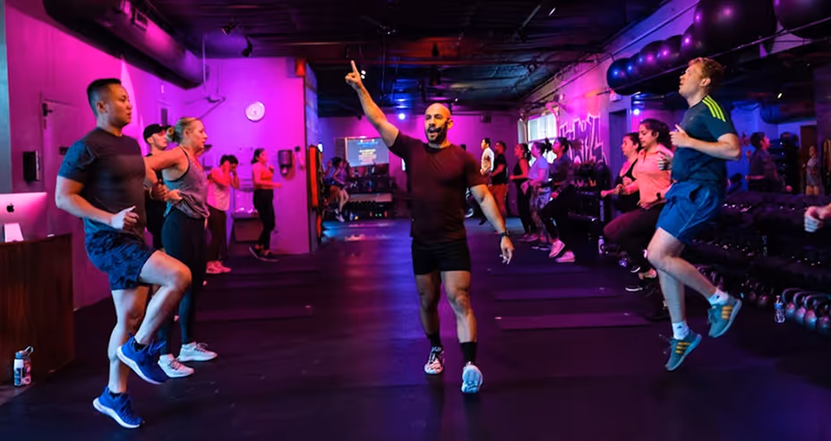 Group fitness class with instructor in the center leading participants through a cardio workout in a dimly lit room with purple and blue lighting.