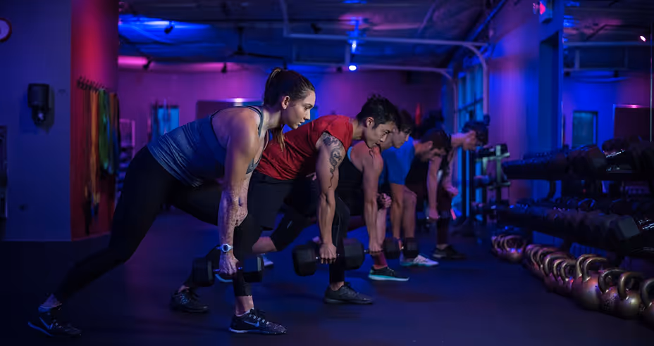 A group of five people in a gym performing bent-over dumbbell rows under purple and blue lighting.