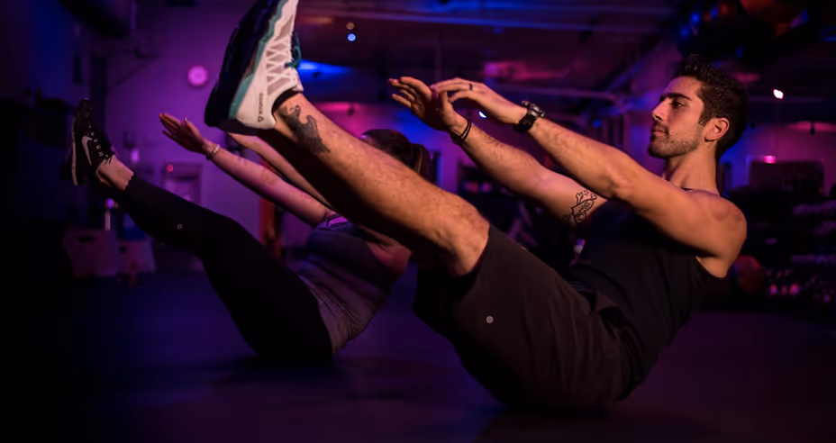 Two people exercising indoors, balancing on their sit bones with legs and arms extended in front in a dimly lit room with purple lighting.