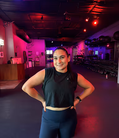 Woman in workout attire standing with hands on hips in a dimly lit gym with red and purple lighting.