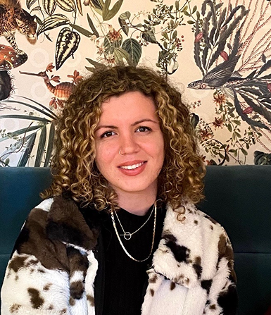 Smiling woman with curly hair wearing a black top and a white and brown spotted coat seated against a jungle-themed wallpaper.