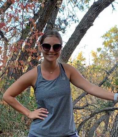 Smiling woman wearing sunglasses and a gray tank top standing outdoors with trees and autumn leaves in the background.