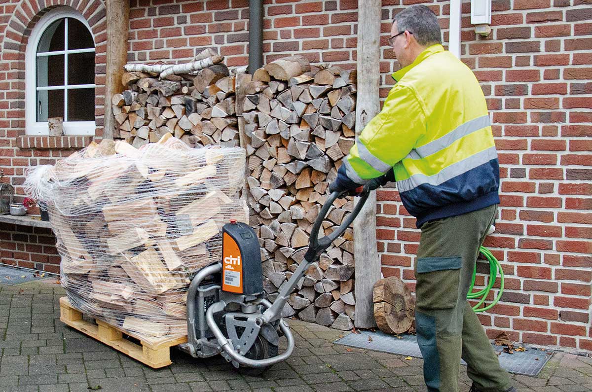 Mann in gelber Jacke bedient elektrischen Handhubwagen mit Holzbündel auf Palette.