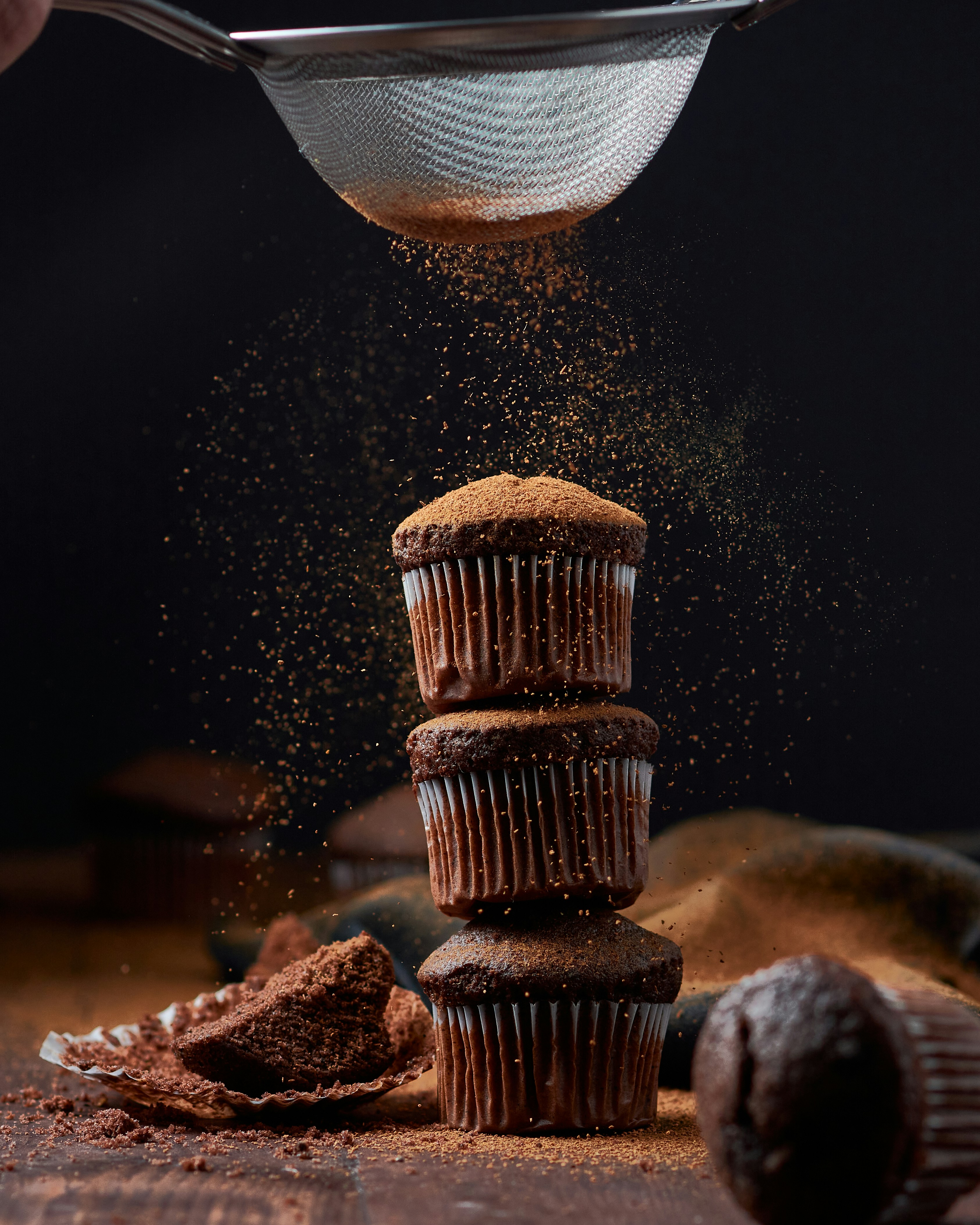 Three chocolate cupcakes stacked in a pile with cocoa being sifted over the top