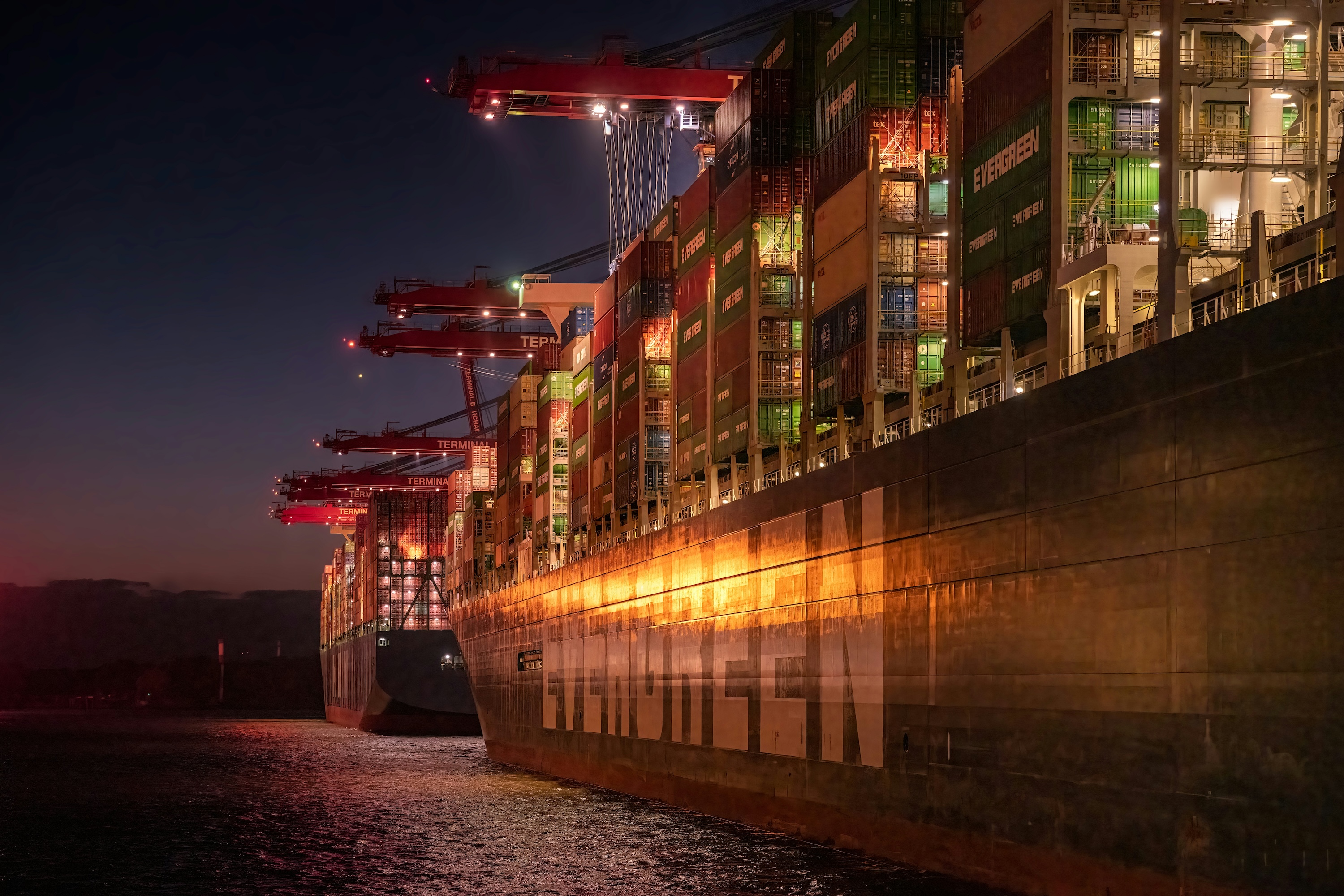 Two docked container ships at night in a lit harbour scene