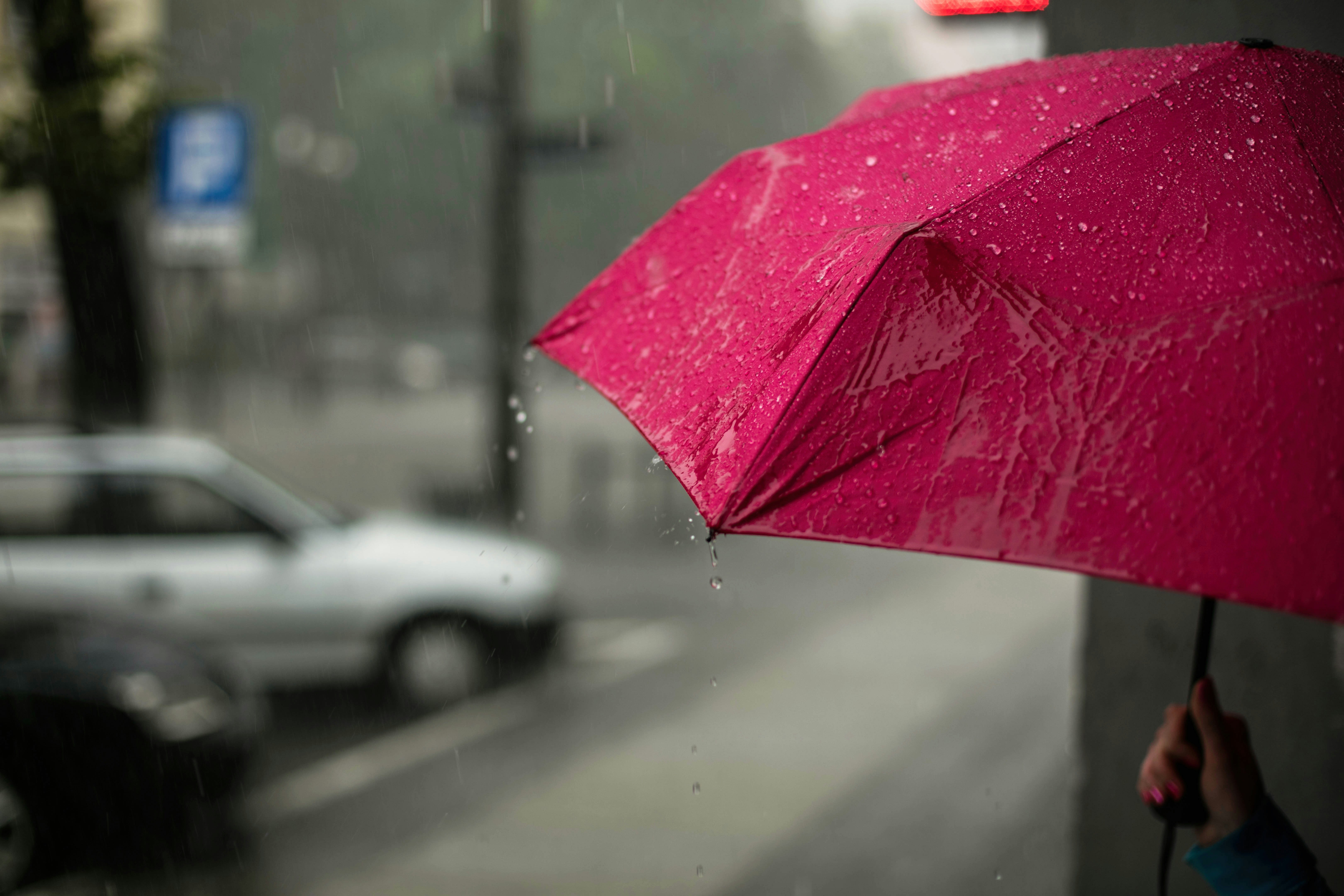 A close up image of a fuschia umbrella held by a womans hand against a drab rainy cityscape