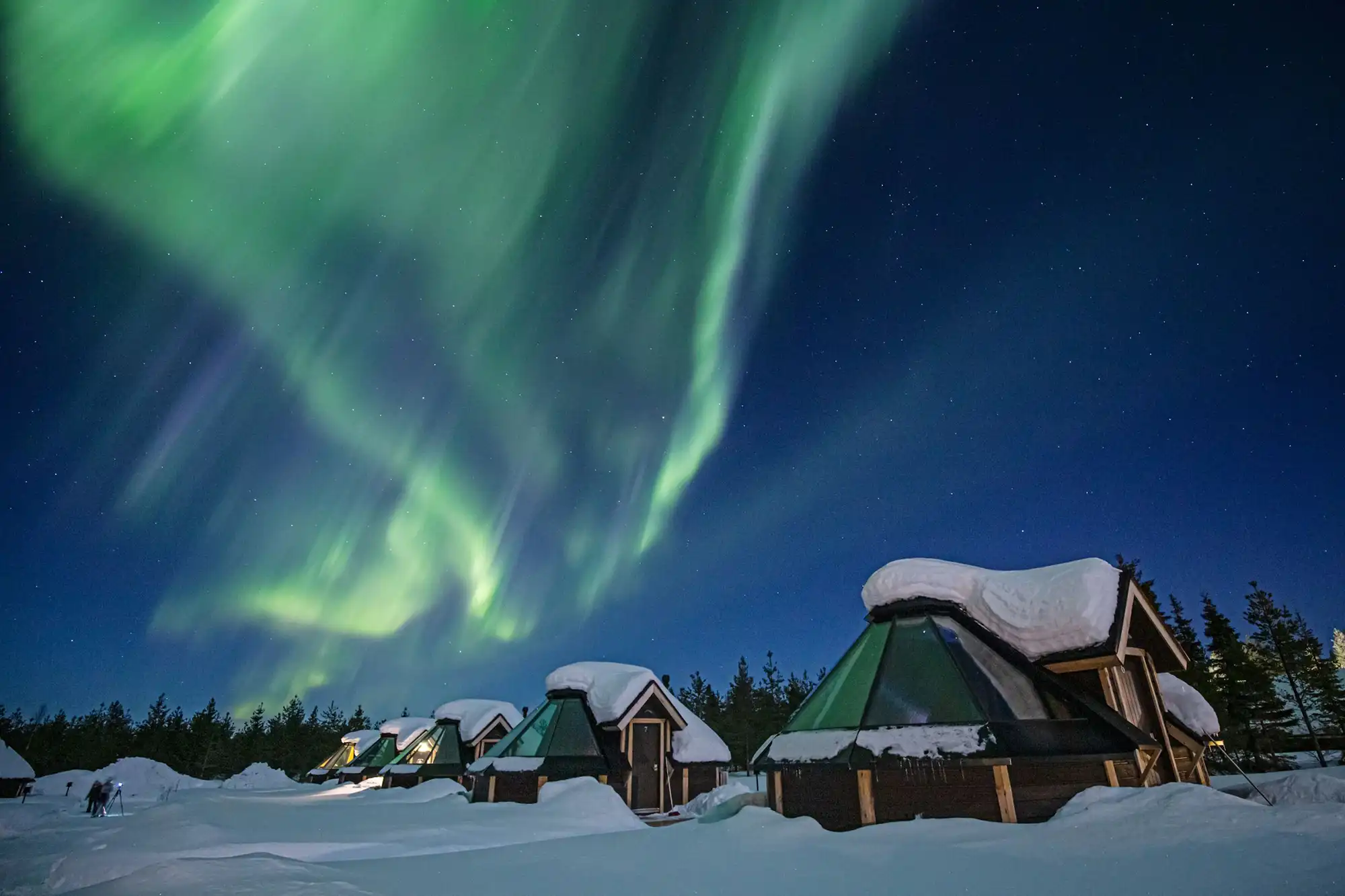 Nordlichter über einer verschneiten Landschaft. Mehrere Hütten mit Glasdach liefern beste Beobachtungsmöglichkeiten im Warmen.