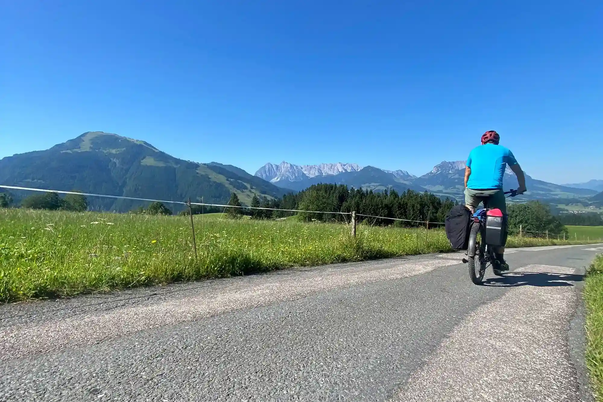Ein Radfahrer auf einem Feldweg zwischen Wiesen, im Hintergrund die Alpen