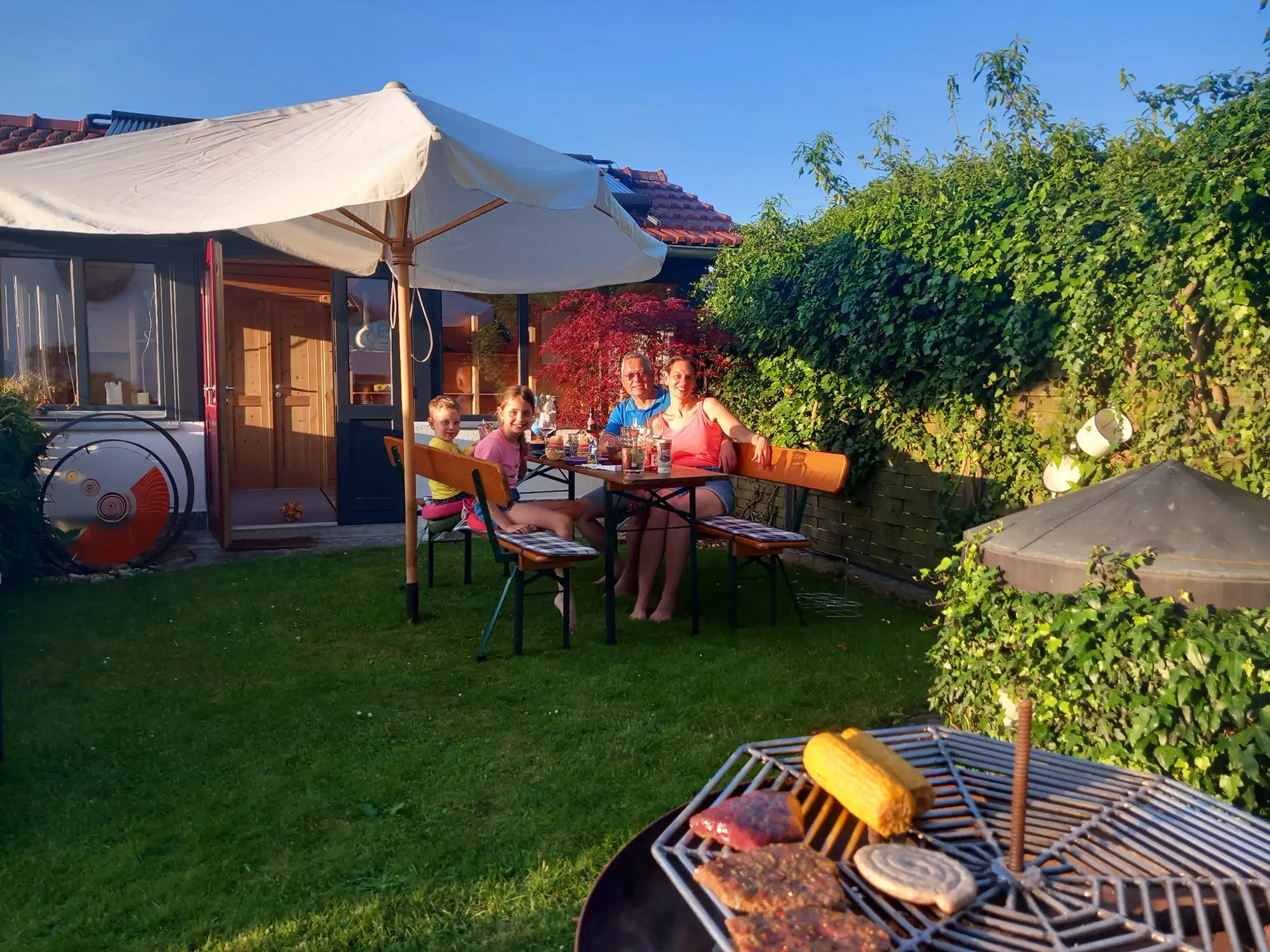 Eine Familie sitzt an einem Tisch im Garten unter einem großen Sonnenschirm, im Vordergrund ein Grill mit Maiskolben und Fleisch.