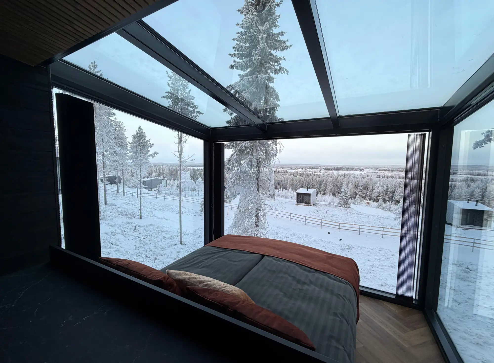 Hochwertige Unterkunft mit Schlafzimmer und Glaswänden und -Glasdach mit Blick auf verschneite Bäume und die Landschaft finnisch Lapplands.