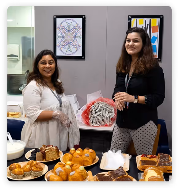 Two women smiling together at a school bake-sale table with assorted pastries