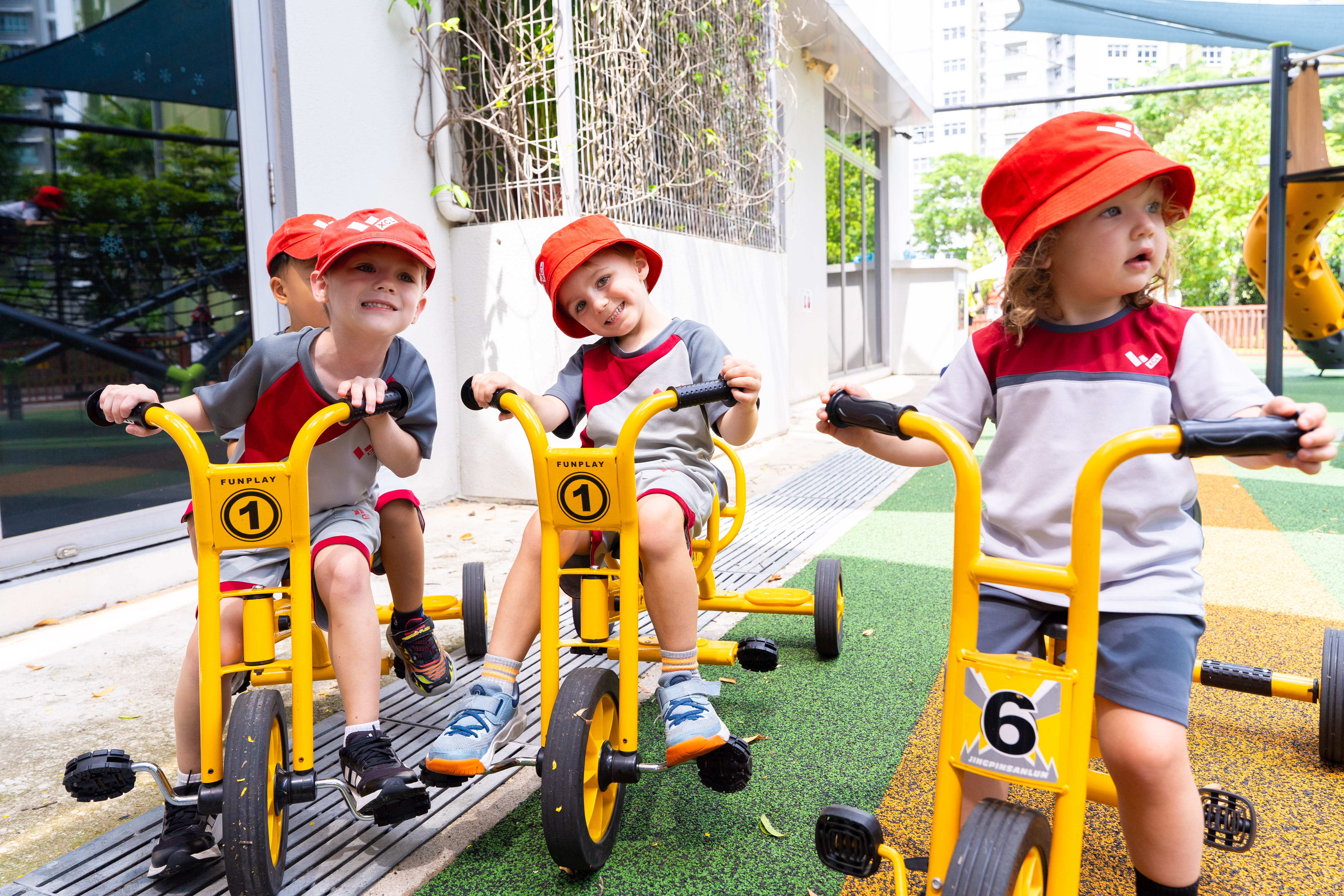 Three young children in XCL uniform riding yellow tricycles on an outdoor playground