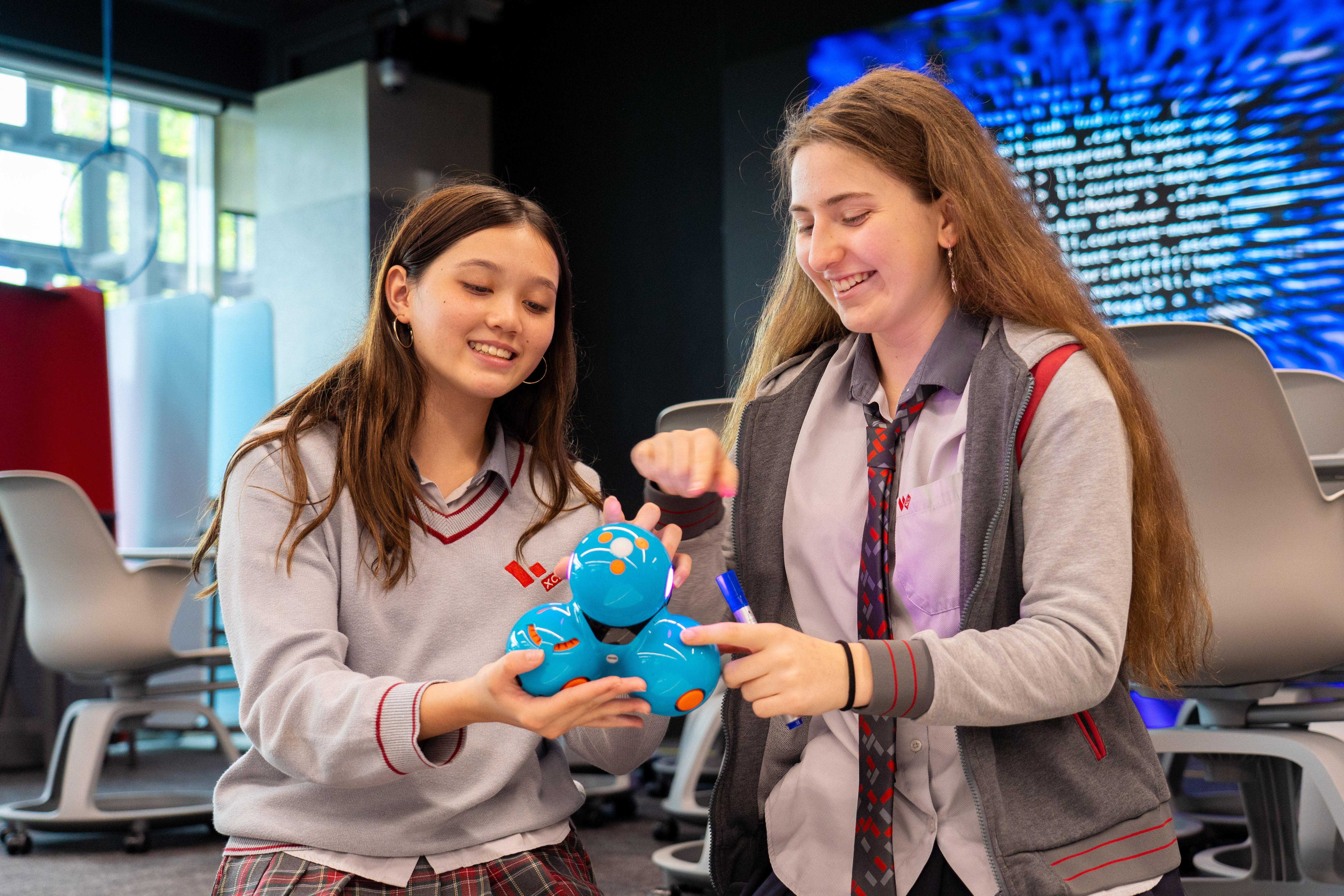 Two students in uniform interact with a blue coding robot in a technology classroom