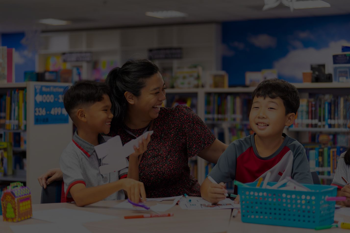 A teacher laughing with two young students at a library table