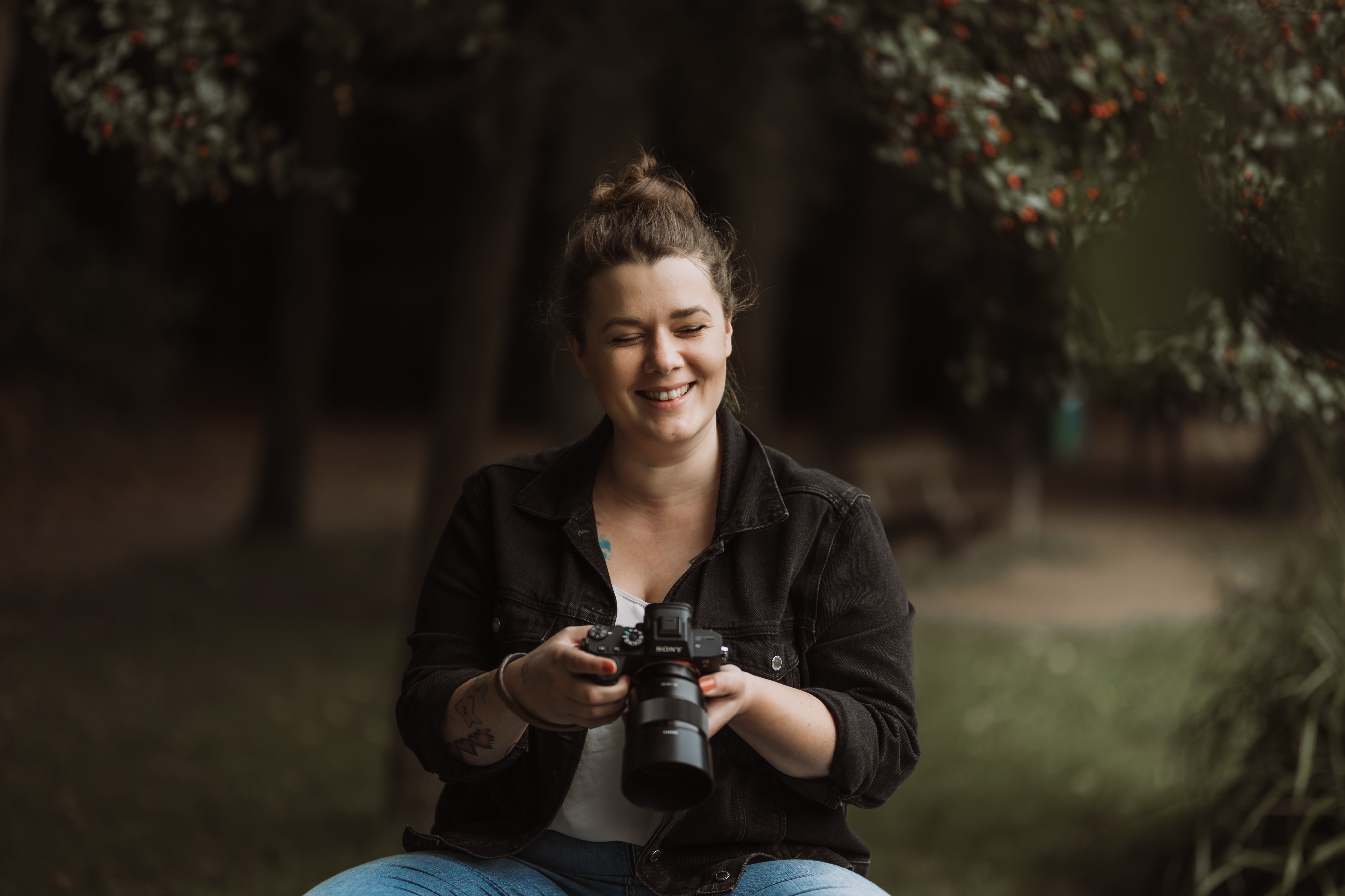 Smiling woman in a black jacket holding a camera outdoors with blurred trees and greenery in the background.