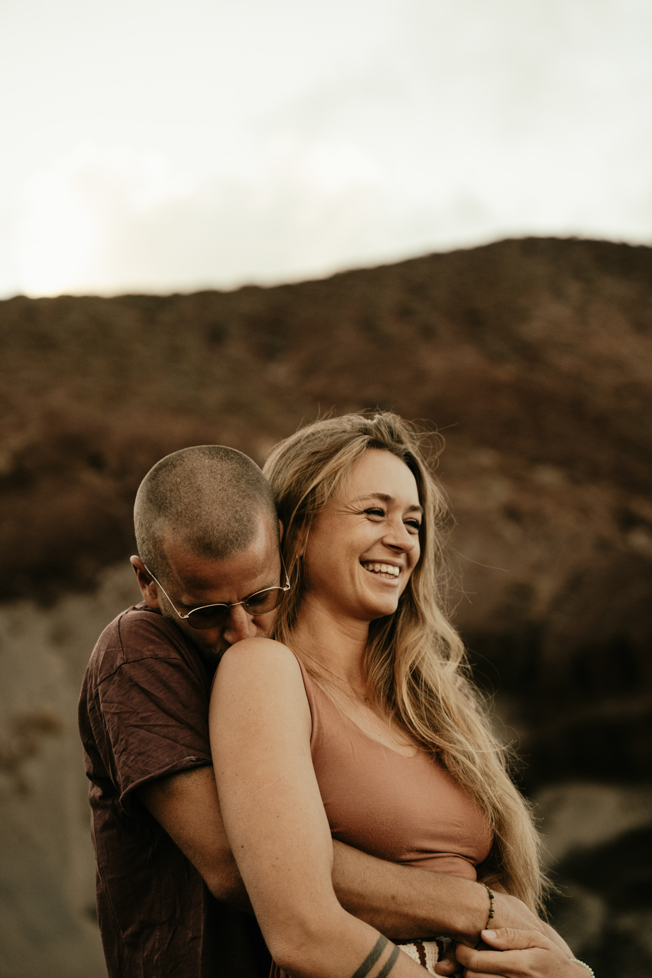 Man with glasses hugging and kissing the shoulder of a smiling woman with long blonde hair against a mountainous background.