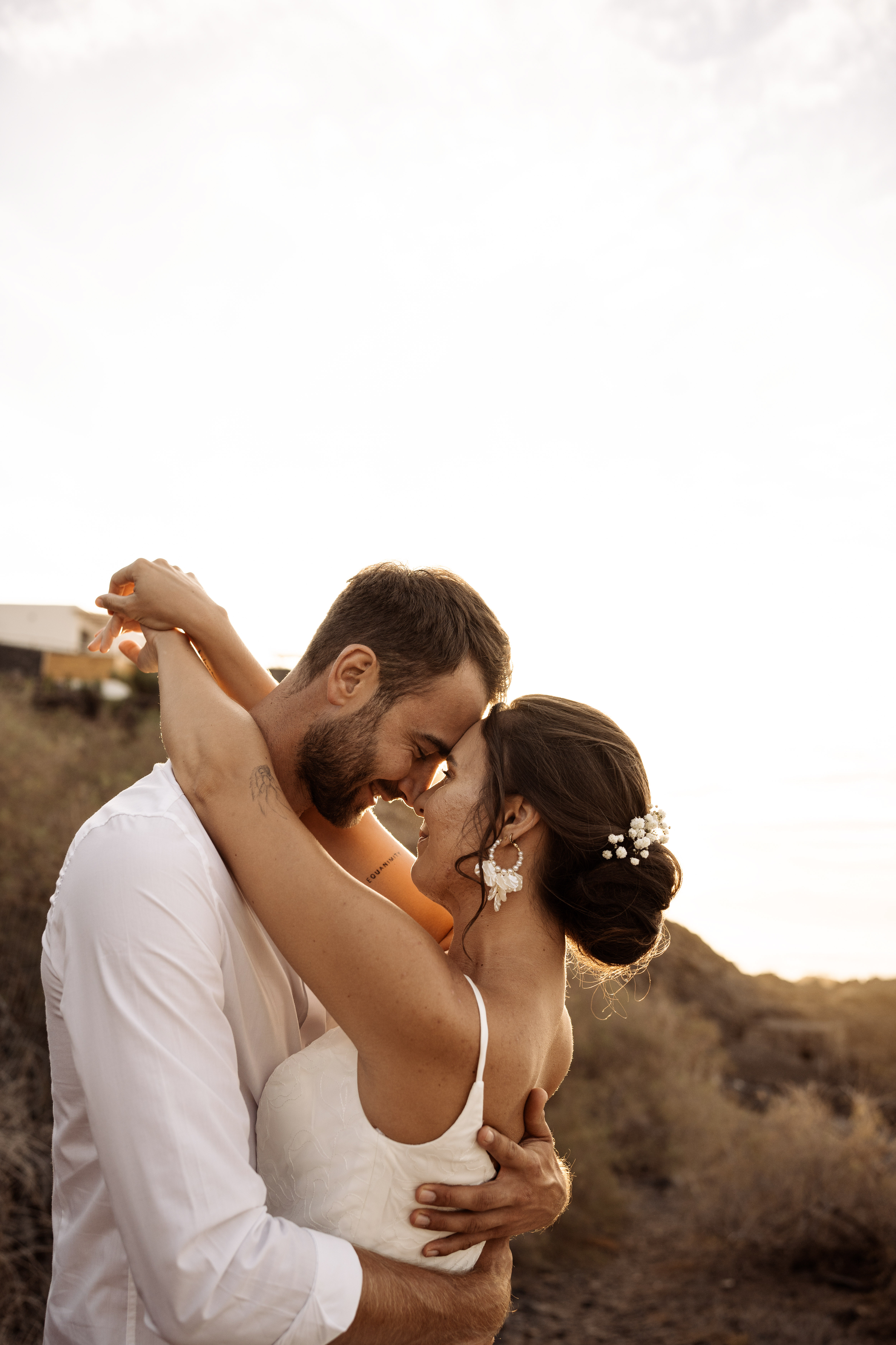 Bride and groom in a loving embrace, touching foreheads outdoors at sunset.