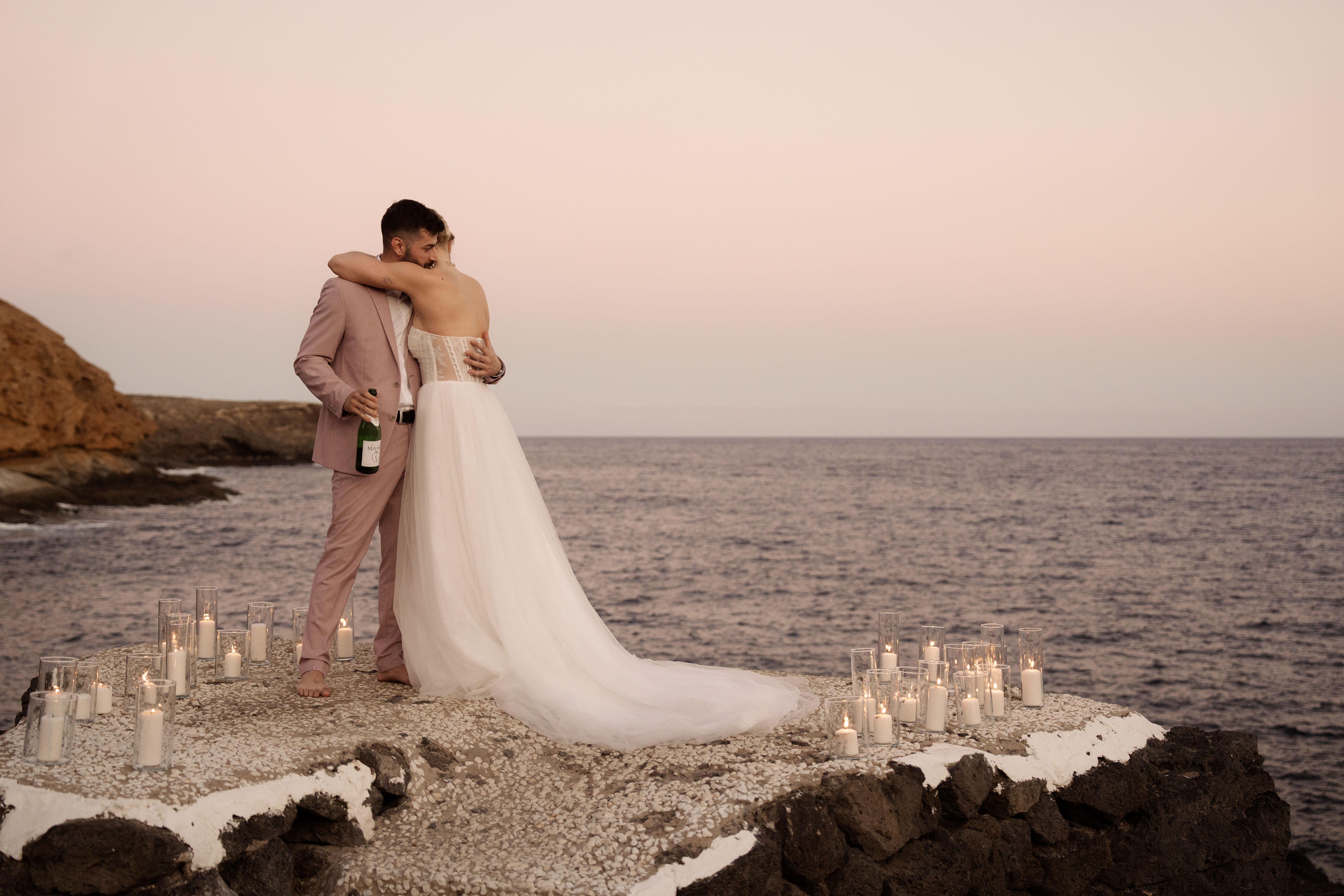 Bride in white wedding dress hugging groom in pink suit holding a bottle, standing barefoot on a rocky ledge by the sea at sunset with lit candles around them.