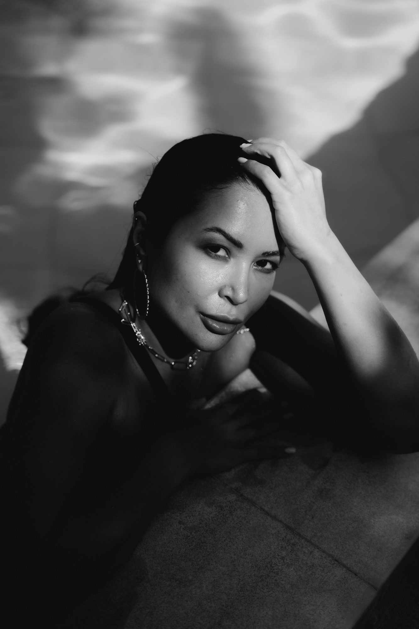 Black and white portrait of a woman resting her arm and hand on a pool edge, looking at the camera with water reflections in the background.